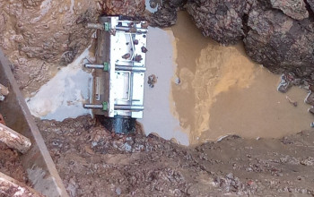 Zdjęcie przedstawia stalowy zacisk montażowy mocujący element rurociągu. Konstrukcja znajduje się w otwartym wykopie wypełnionym błotem i wodą gruntową.