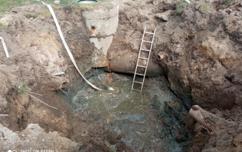 Zdjęcie przedstawia otwarty wykop ziemny, w którym trwają prace związane z wymianą elementów przepompowni ścieków. Widać betonową studnię, do której doprowadzony jest stary rurociąg, częściowo zanurzony w wodzie gruntowej i błocie.