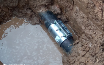 To zdjęcie przedstawia grubą, czarną rurę częściowo zanurzoną w błotnistej wodzie, wewnątrz wykopu ziemnego. Na środku rury widoczny jest metalowy pierścień zaciskowy z prętami i śrubami.