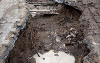 To zdjęcie przedstawia duży, głęboki wykop w ziemi, który odsłania glebę i gruz. Na dnie widoczna jest jasna kałuża płynu, a w górnej części zakopane rury i cegły.