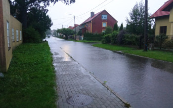 Woda pokrywa całą szerokość jezdni, sięgając aż do krawężnika przy chodniku z kostki brukowej, na którym widoczna jest pokrywa studzienki.