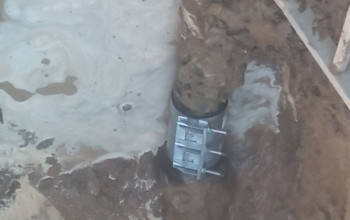 Metalowa opaska naprawcza jest szczelnie zamontowana na skorodowanej rurze żeliwnej, znajdującej się w wykopie ziemnym wypełnionym błotnistą wodą. 